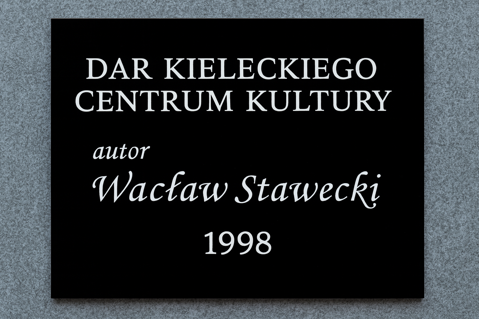 Tablica pamiątkowa z czarnej plexi z białym napisem: Dar Kieleckiego Centrum Kultury, autor Wacław Stawecki, 1998